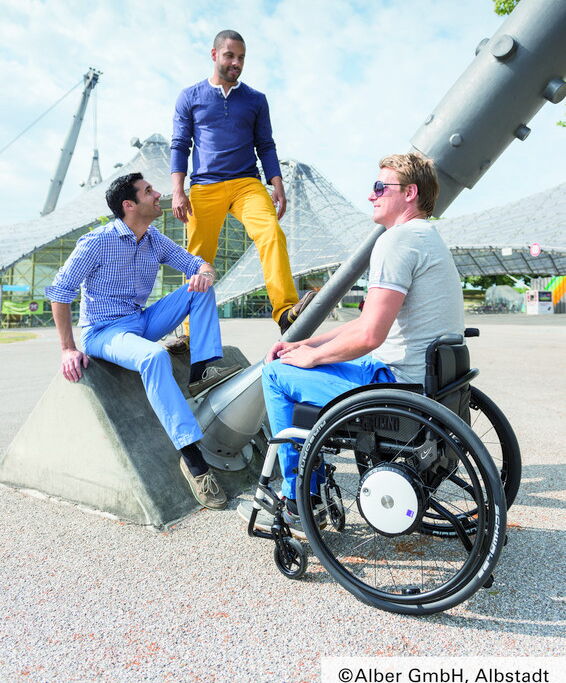 3 Personen auf dem Olympiagelände in München, eine Person im Aktivrollstuhl